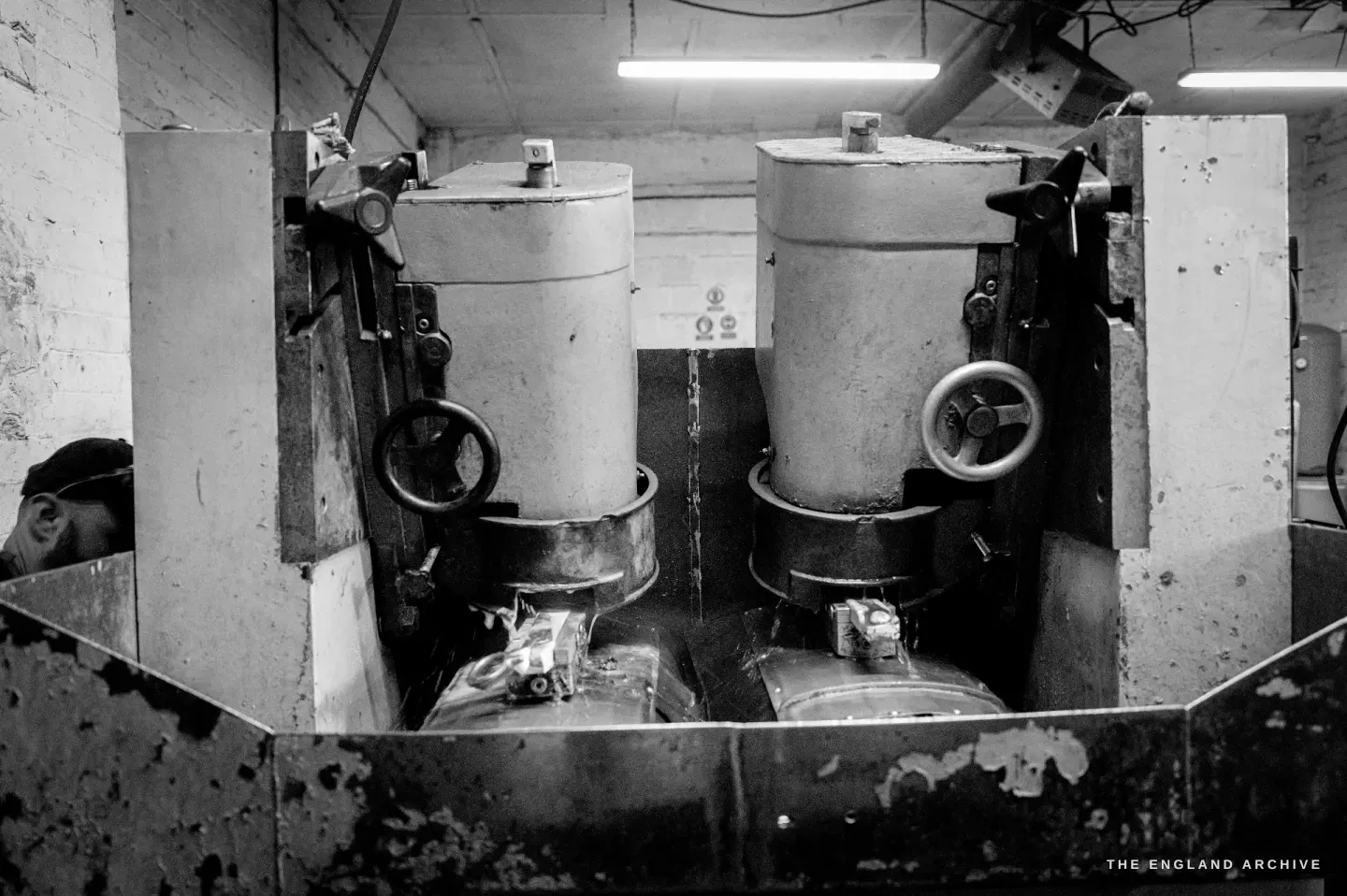 Two grinding machines head-on - tall cylindrical housings over abrasive wheels, with troughs beneath, set into a worn bench in the dark of the workshop.