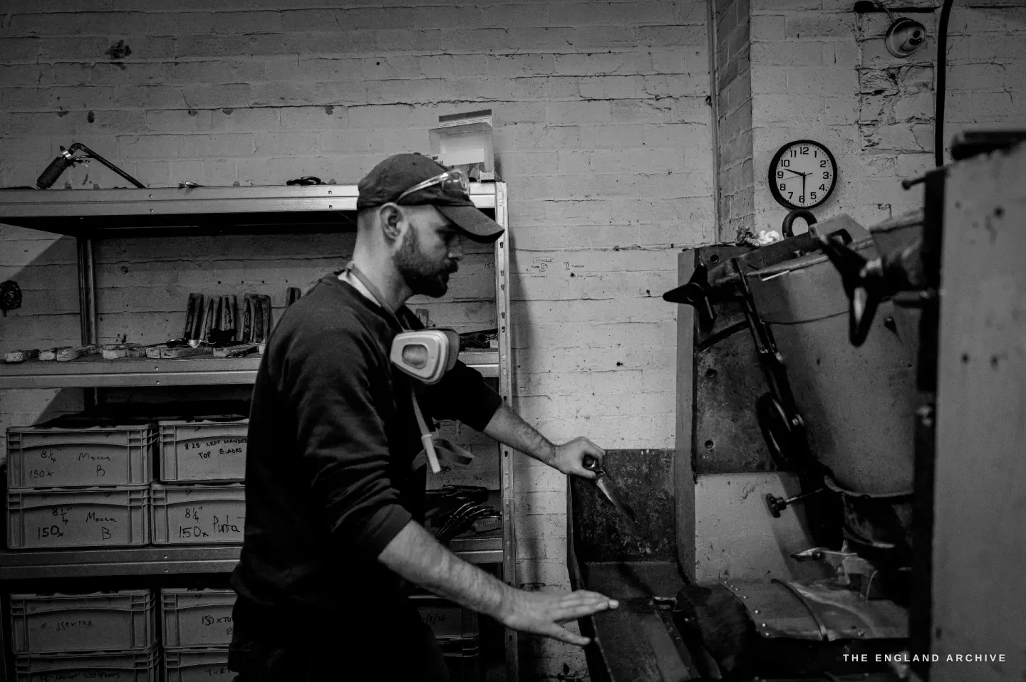 Jonathan at the grinding machine, ear defenders round his neck, a blade in one hand, stacked bins of parts labelled by model and size beside him.