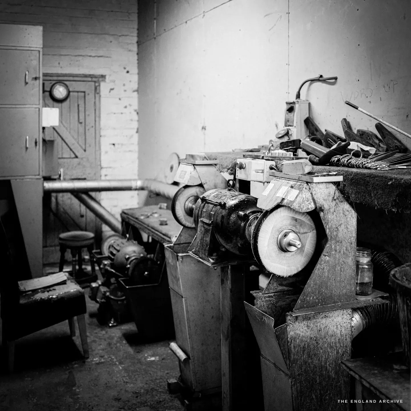 A glazing station: belt-driven polishing wheels on a spindle, brushes and tools racked behind, a stool and a doorway beyond.