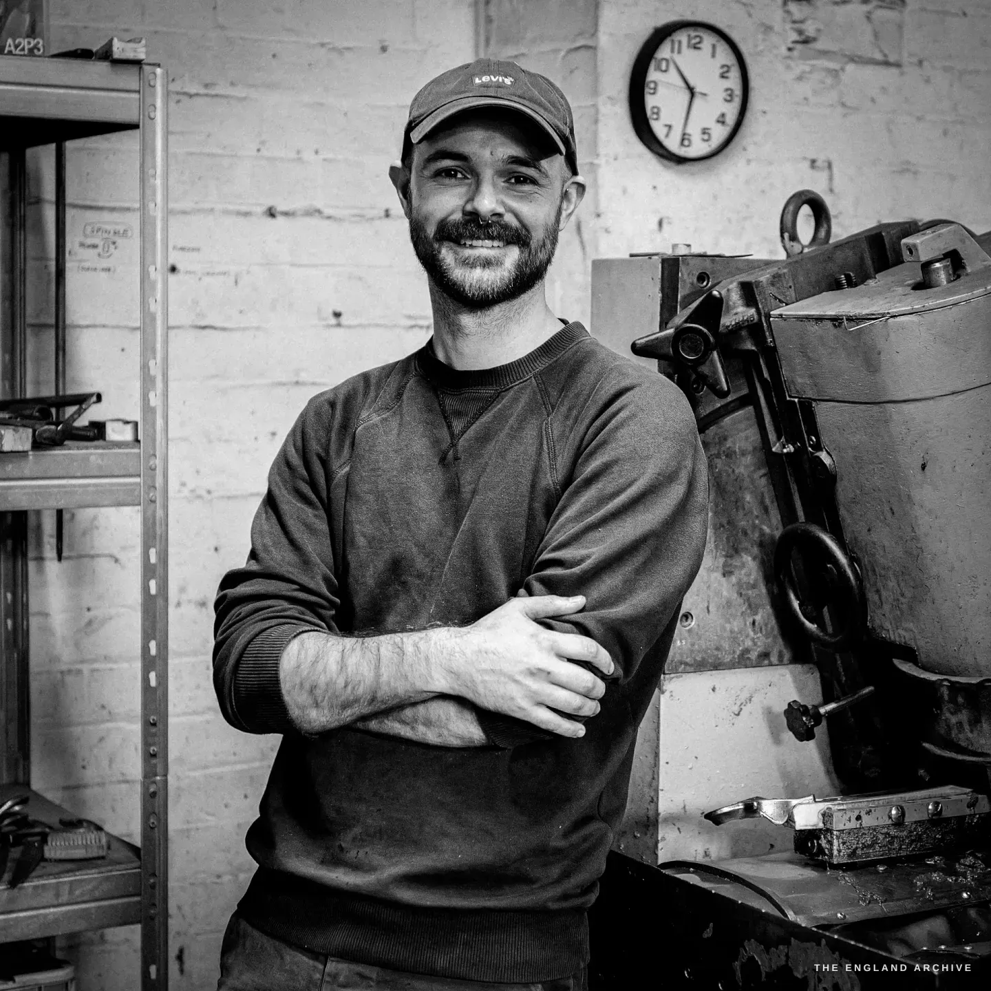 A square portrait of Jonathan, arms folded, smiling, the wall clock above his shoulder and the grinding machine to the right.