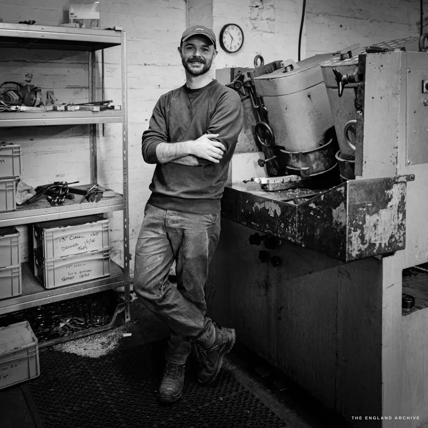 A full-length portrait of Jonathan leaning against the grinding machine, arms folded, one foot crossed, the clock and shelves behind him.