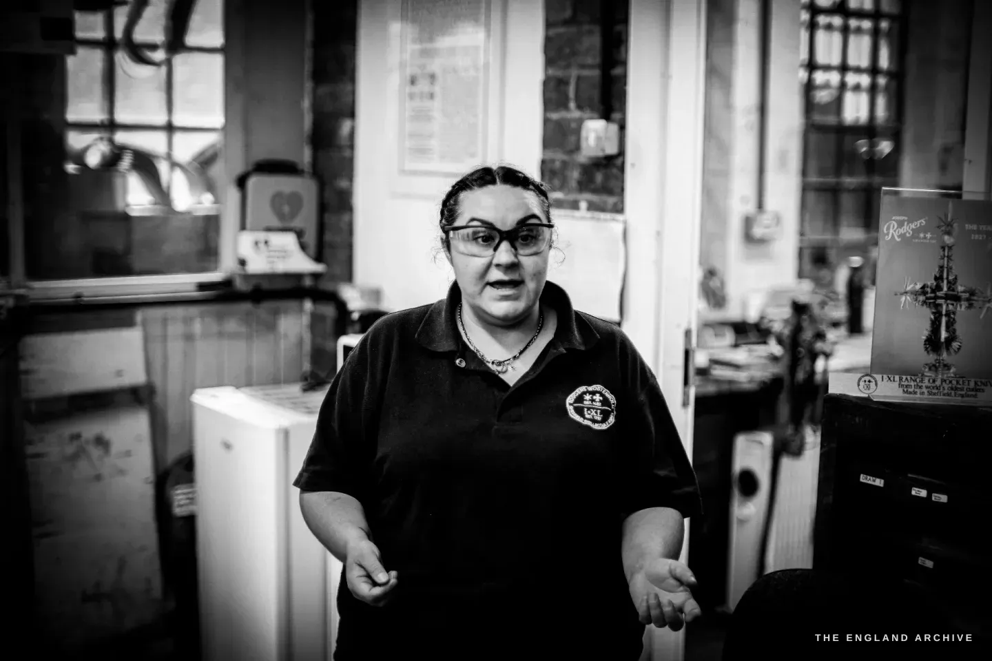 Kylie Cocker stands facing the camera in the workshop, hands open at her sides, in a dark Rodgers Wostenholm polo shirt, with industrial machinery and a framed cross behind her.