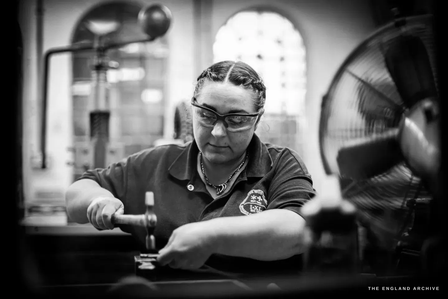 Kylie Cocker leans over her bench tapping a small knife part on a steel stake with a light hammer, safety glasses on, an arched window and a fan behind her.