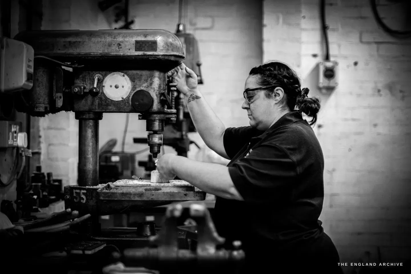 Kylie Cocker works a cast-iron pillar drill, lowering the bit onto a small component clamped to the table, safety glasses on.