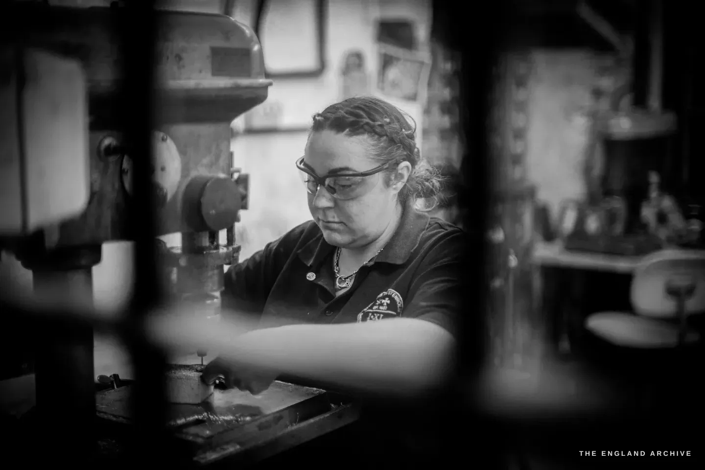 Framed by the column and head of an old drill press, Kylie Cocker works at the machine, her Rodgers Wostenholm badge and the workshop&rsquo;s framed pictures visible behind.