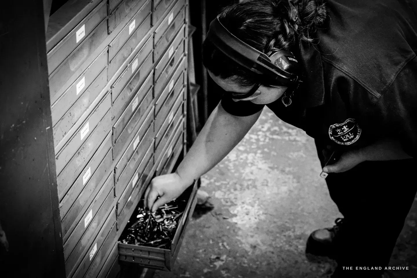 Kylie Cocker bends to a tray of small steel components set on the floor in front of a tall bank of labelled wooden parts-drawers.
