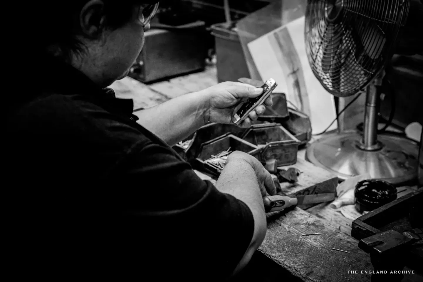 Kylie Cocker works a small knife in both hands at her bench, a fan and scattered parts beside her.