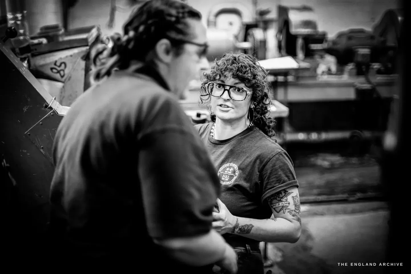 Kylie Cocker, her back to the camera, talks with her colleague Emma Lawes, who listens in glasses and a t-shirt with a tattooed forearm, machines behind them.