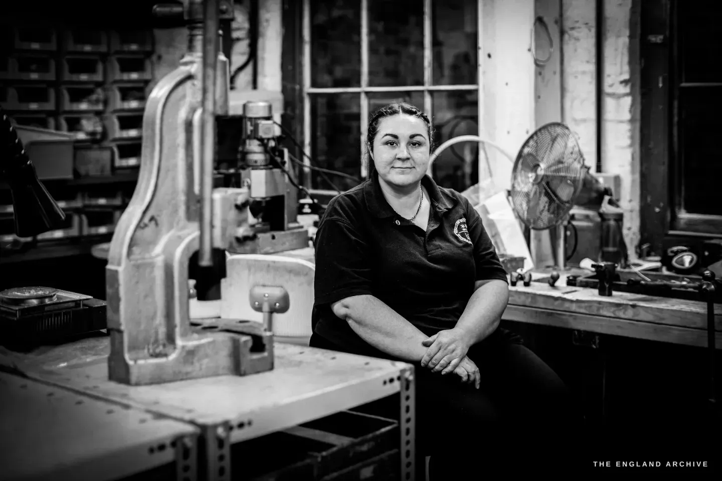 Kylie Cocker sits at her bench facing the camera, hands folded, a fly press and a fan around her in the workshop.