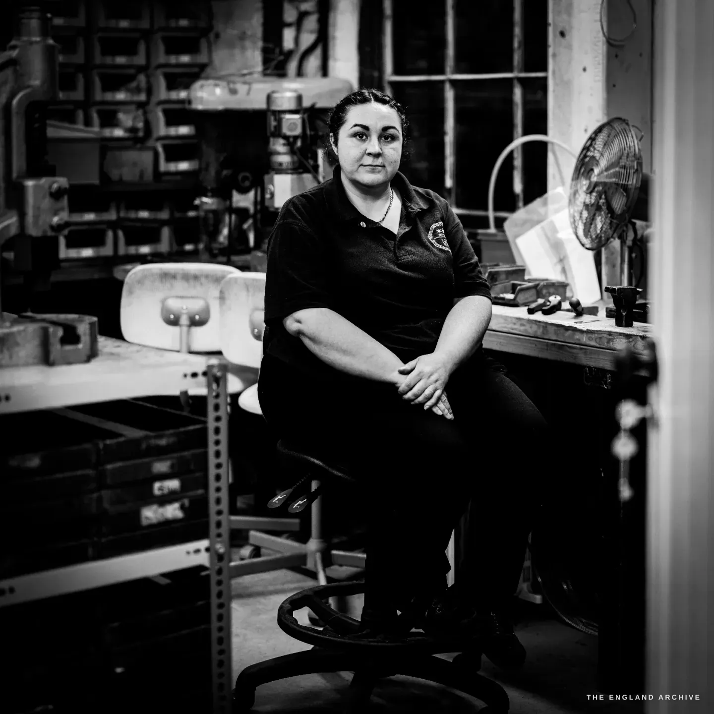 Kylie Cocker sits on her work stool facing the camera, the bank of parts-drawers to one side and the museum&rsquo;s arched windows behind.