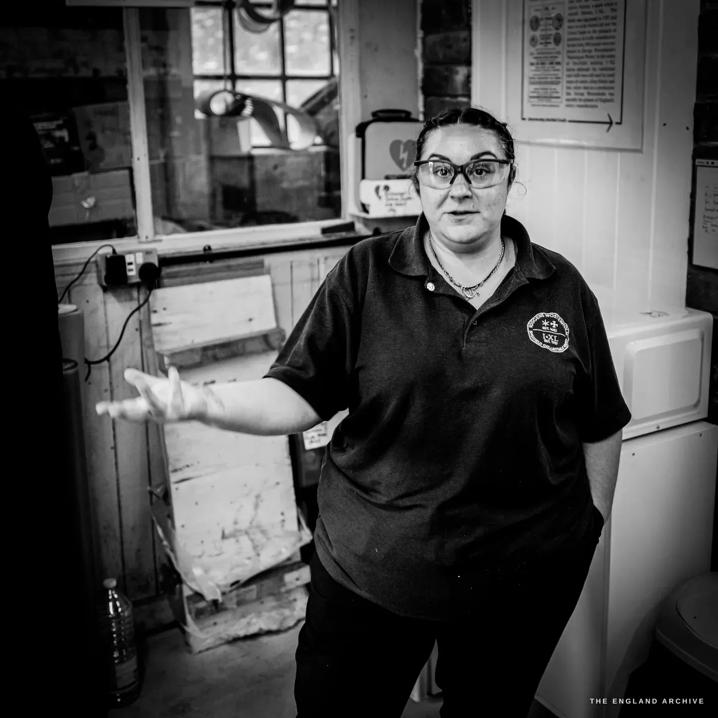 Kylie Cocker stands in the workshop mid-explanation, one hand raised, beneath notices and an arched window.