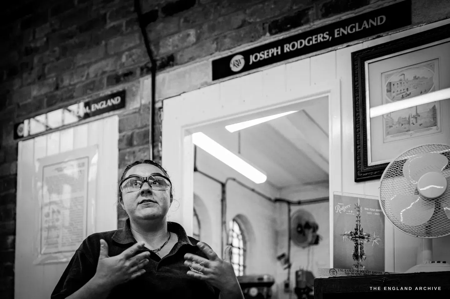 Kylie Cocker stands talking beneath a painted sign reading Joseph Rodgers, England, with the firm&rsquo;s star and Maltese cross mark and framed prints around her.