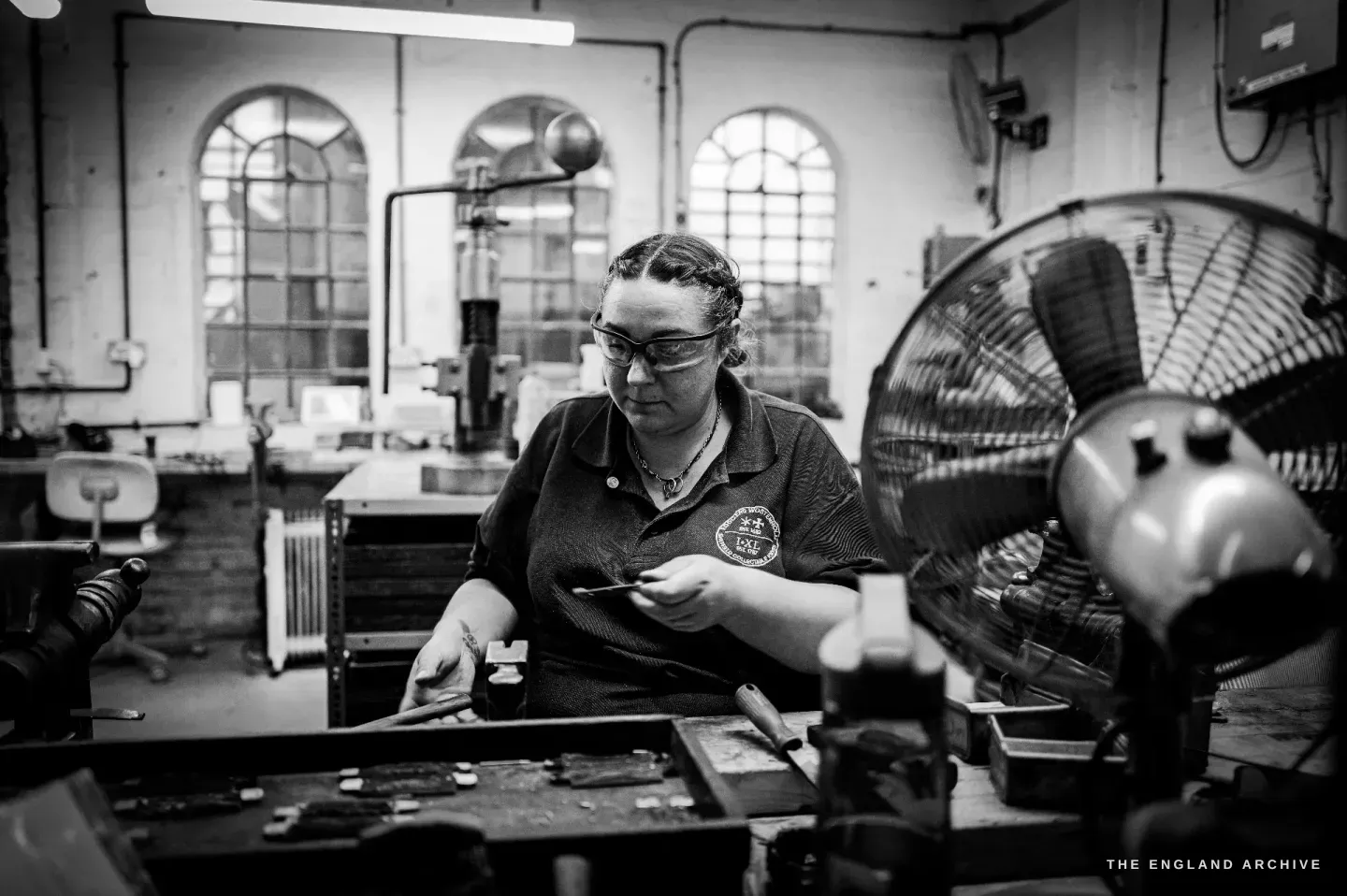 Kylie Cocker works at a long bench beneath the tall arched windows of the Kelham Island workshop, a large fan beside her and trays of parts in front.