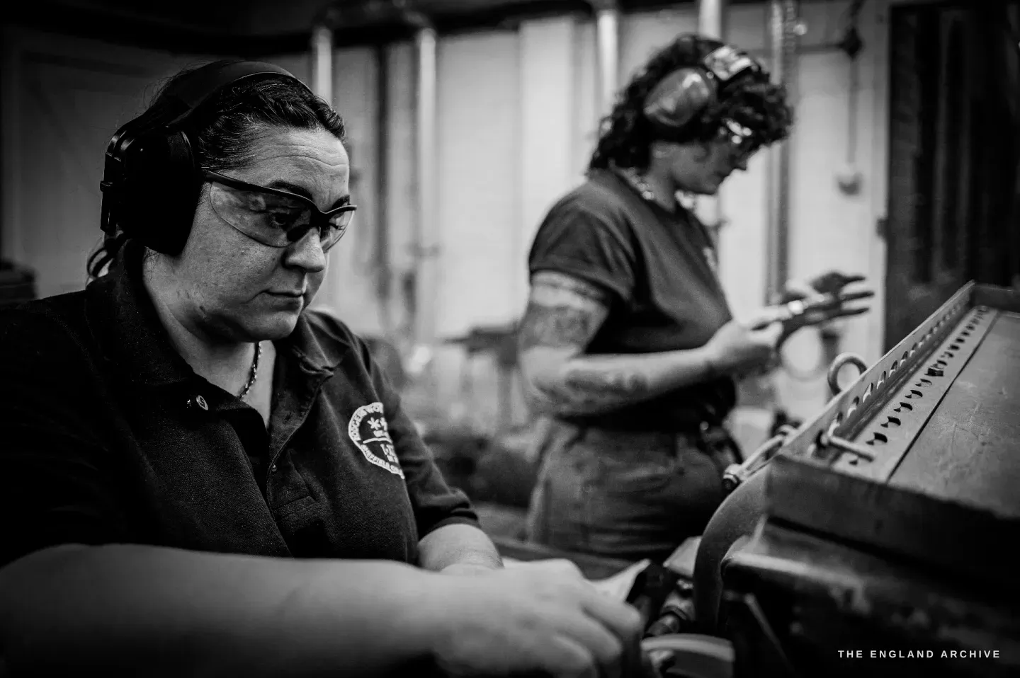 Kylie Cocker works in the foreground in ear defenders while her colleague Emma Lawes works at a machine behind her, both in the workshop.