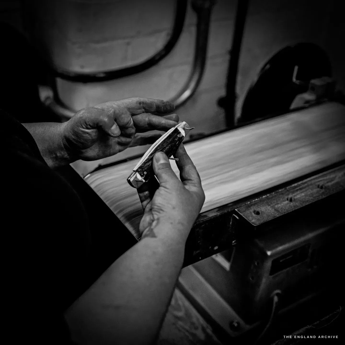 Hands hold a pocket knife against the flat moving belt of a linisher, finishing the edge of the scales.
