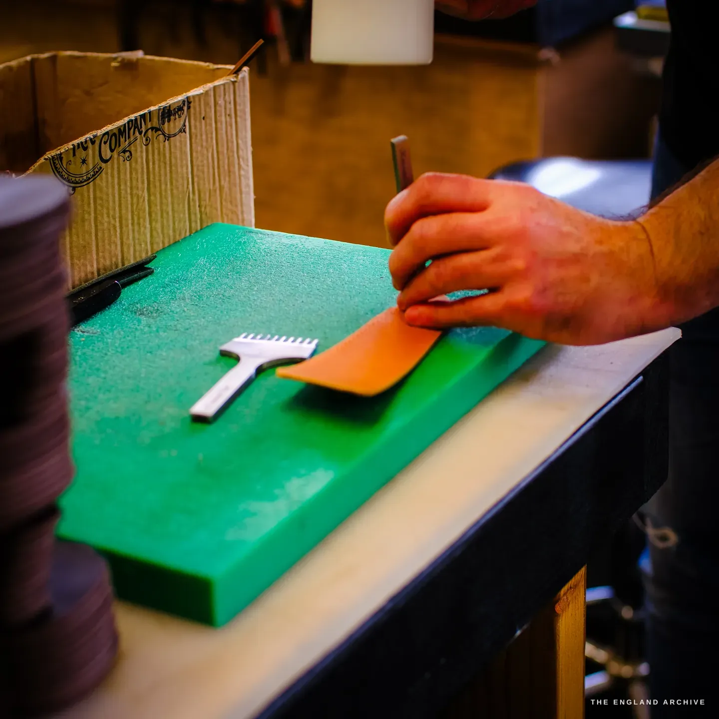A hand draws a stick of edge dressing along the curved edge of a tan leather strip on a green cutting board, a metal comb tool beside it.