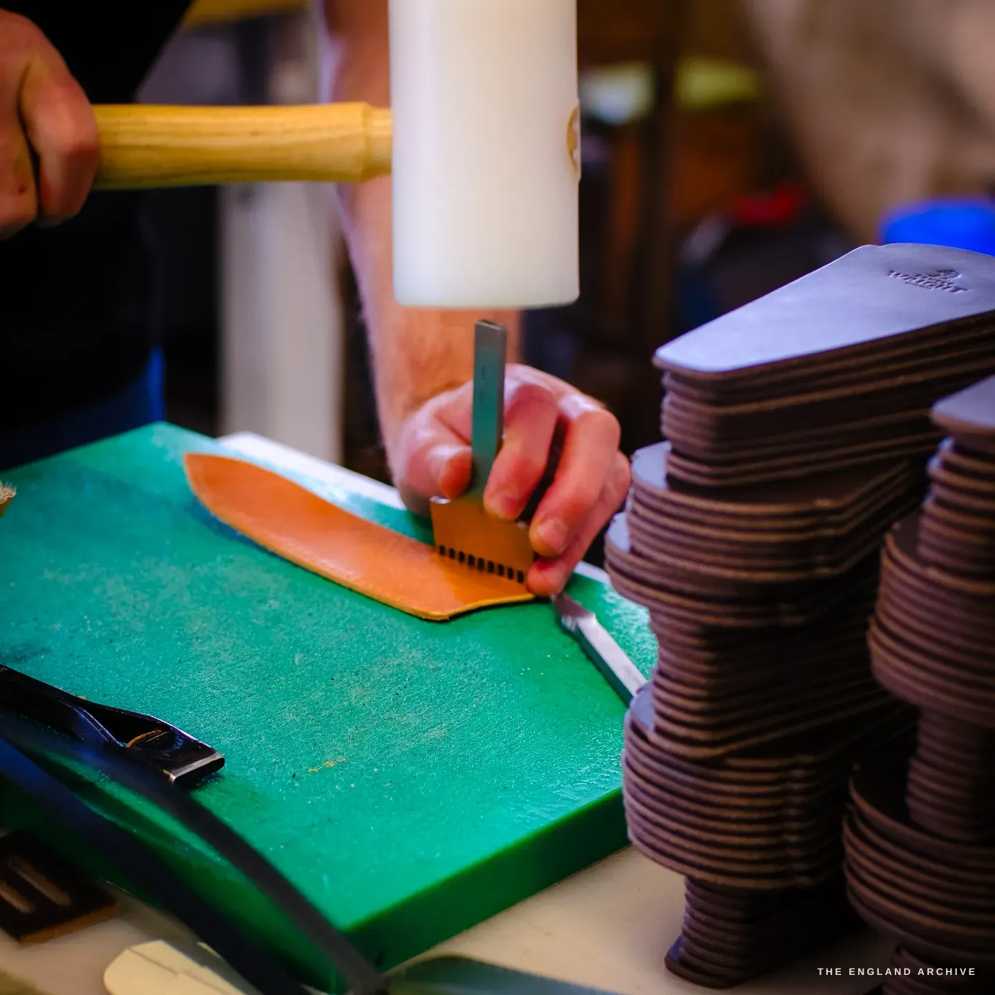 A mallet drives a stitching iron through a tan leather pouch on a green board, with stacks of cut leather blanks beside it.