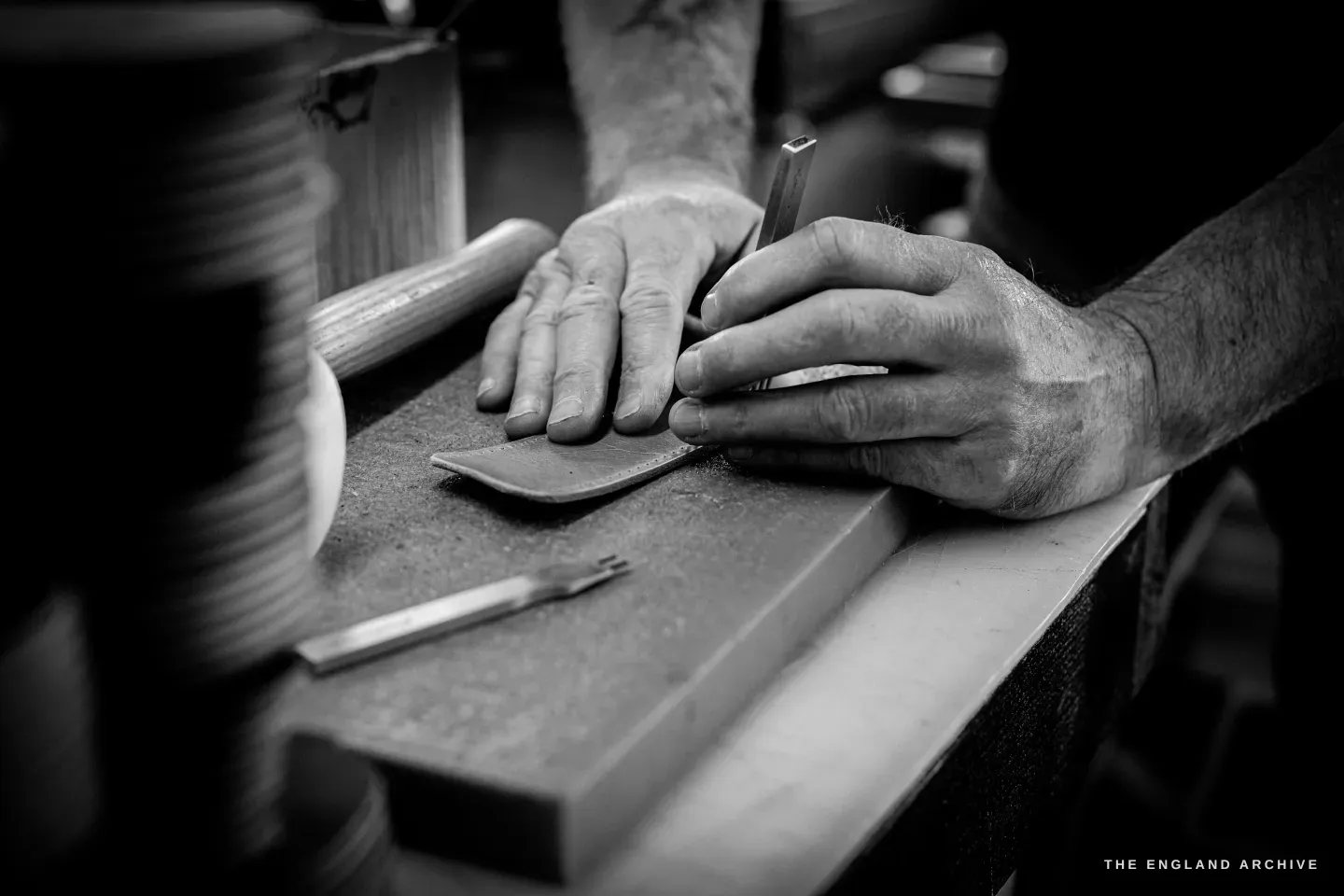 Close on Kevin Wilebore&rsquo;s hands marking a small curved piece of leather with a creaser on the bench, a bobbin of thread to the side.