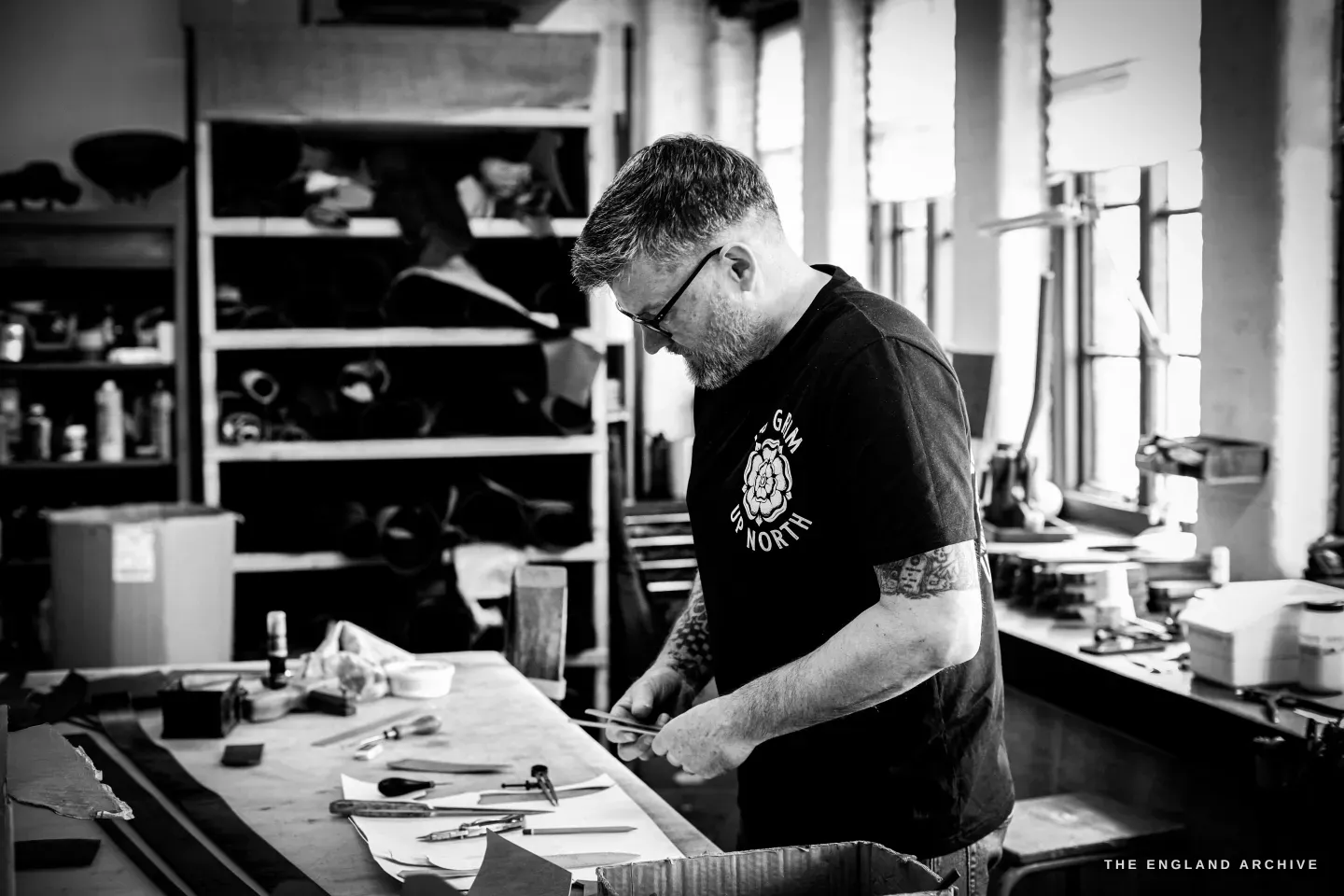 Kevin Wilebore works at his long bench in the airy workshop, tools laid out, shelves of leather behind and bright windows to the right.