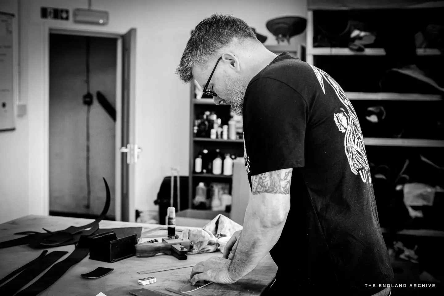 Kevin Wilebore bent over his bench working, shelves of dye bottles and offcuts around him and a fire-exit door to the left.