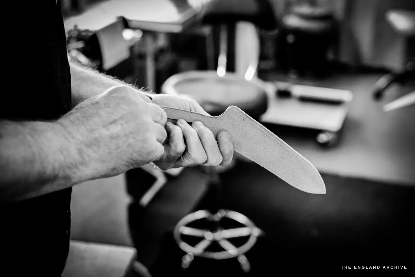 Kevin Wilebore holds a long curved leather blank up in both hands to check it, a workshop stool behind.