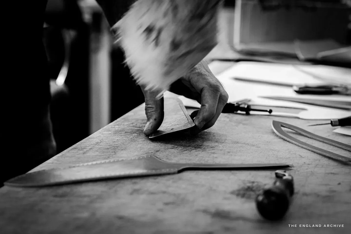 A hand burnishes the edge of a long leather strap on the bench, the movement blurred, dividers and a strip of leather alongside.