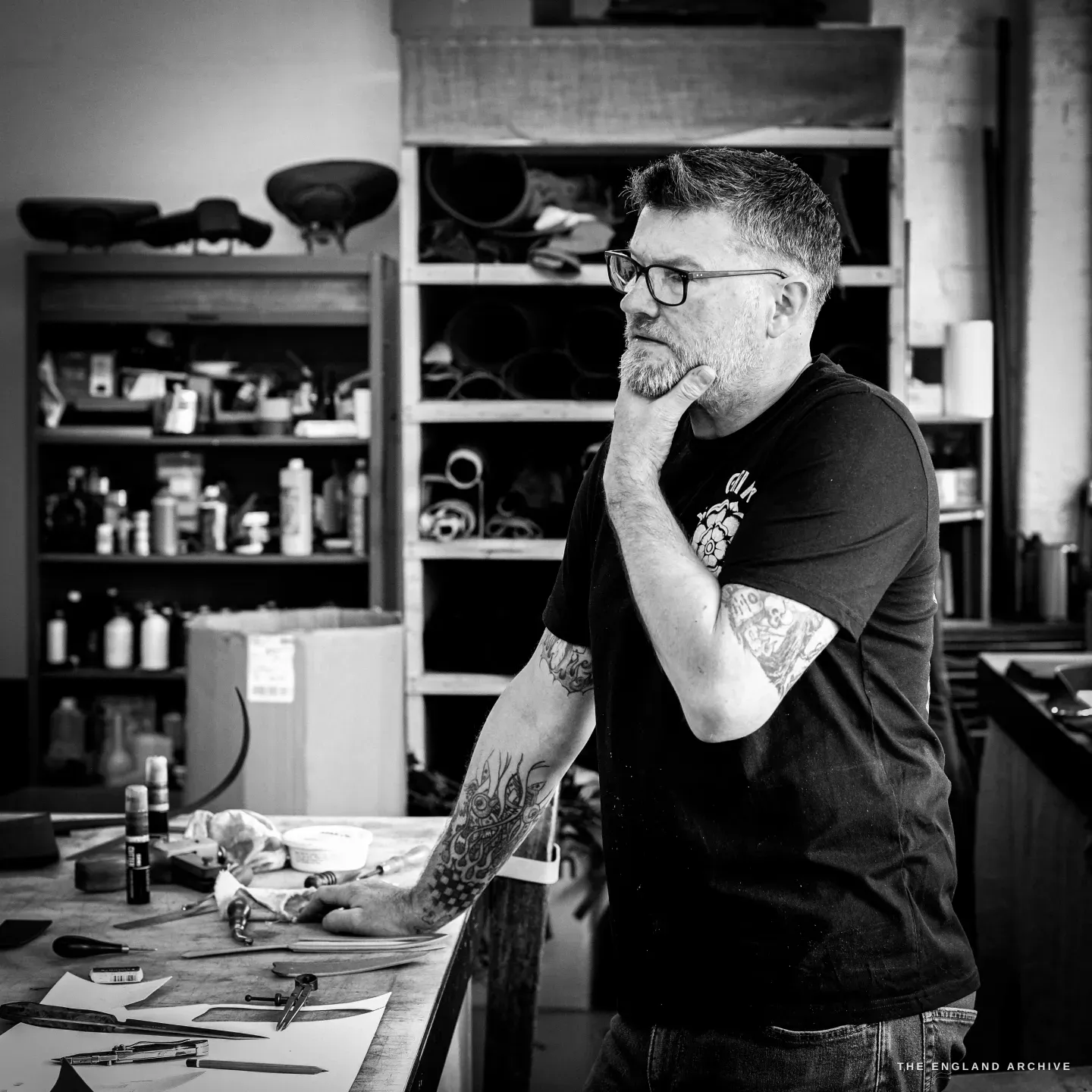 Kevin Wilebore stands with a hand to his beard, thinking, in front of shelves of dye bottles and rolled hides.