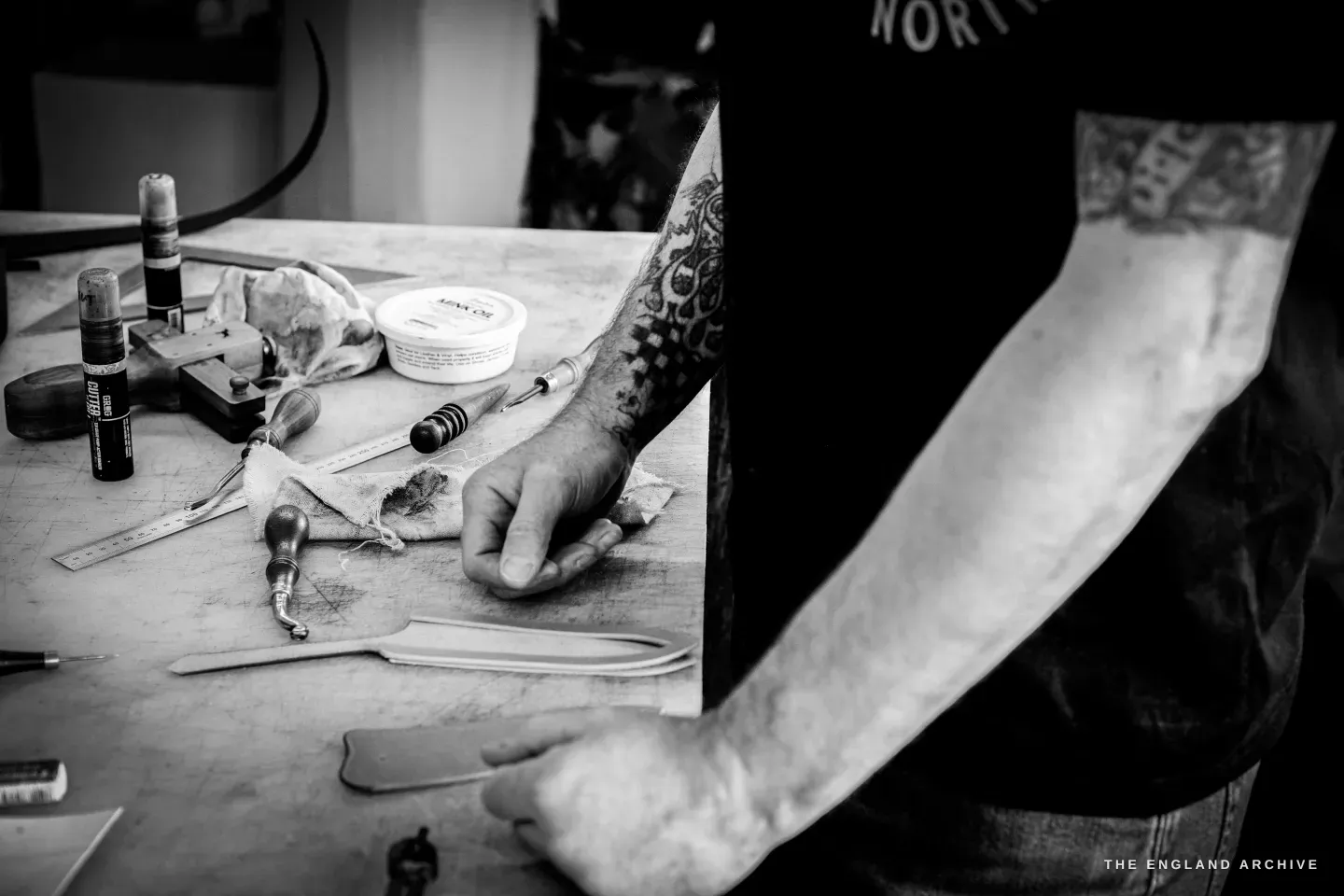 Close on Kevin Wilebore&rsquo;s tattooed arms and a row of leather tools on the bench as he works a pale strip.