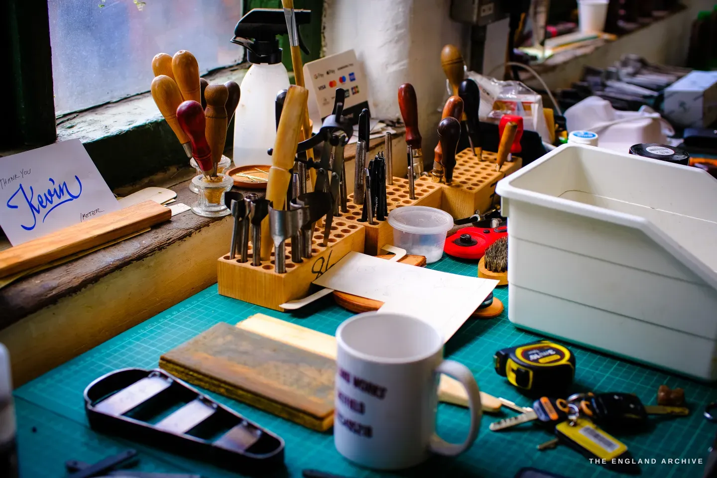Kevin Wilebore&rsquo;s workbench: a rack of leather hand-tools, a green cutting mat, a mug and a card reading Kevin.