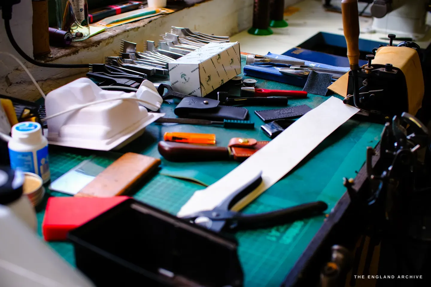 A bench scattered with leather tools, a box of metal fittings, knives and a leather phone case on a green mat.