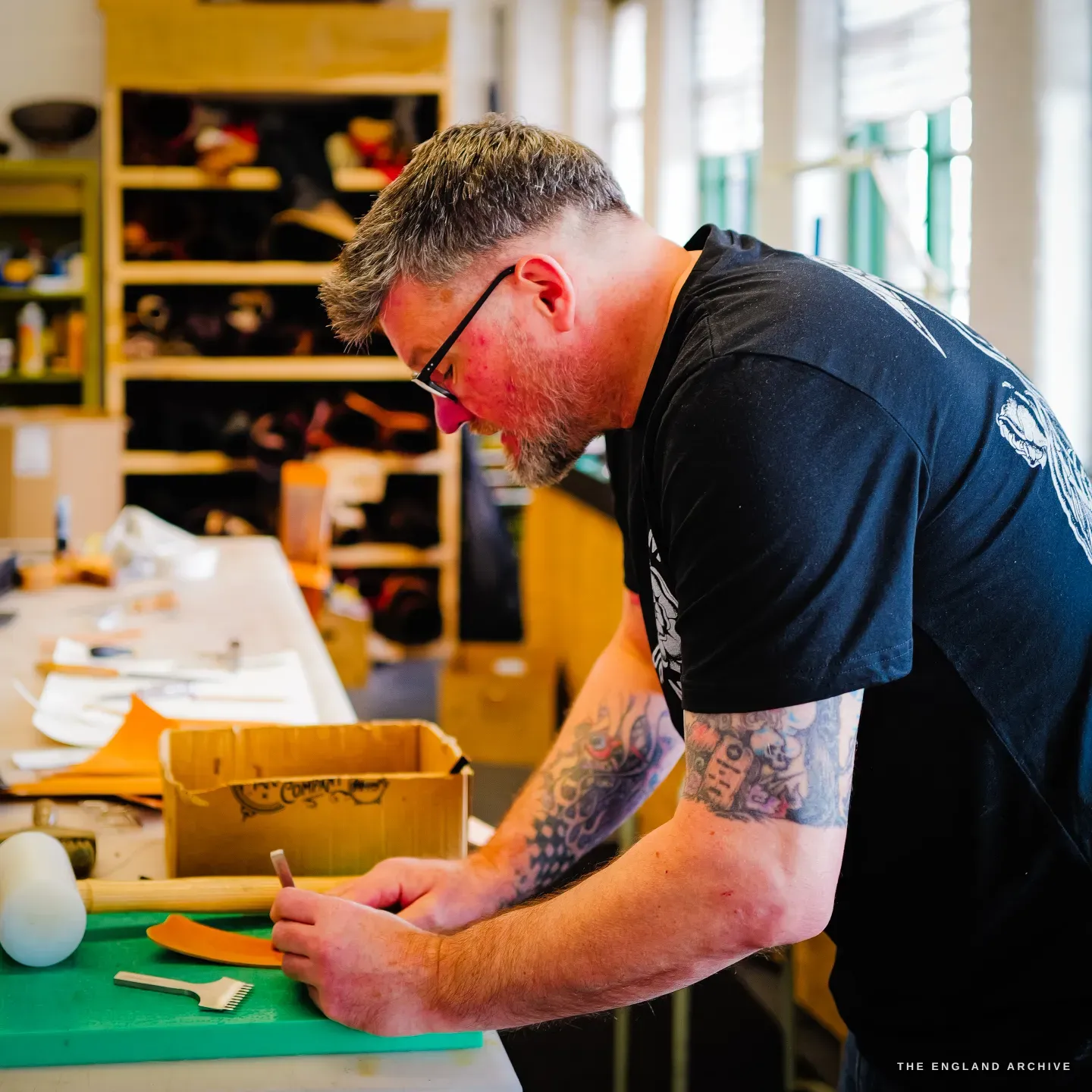 Kevin Wilebore in profile marking a tan leather piece at the bench, the bright bench-lined workshop and shelves of goods behind.