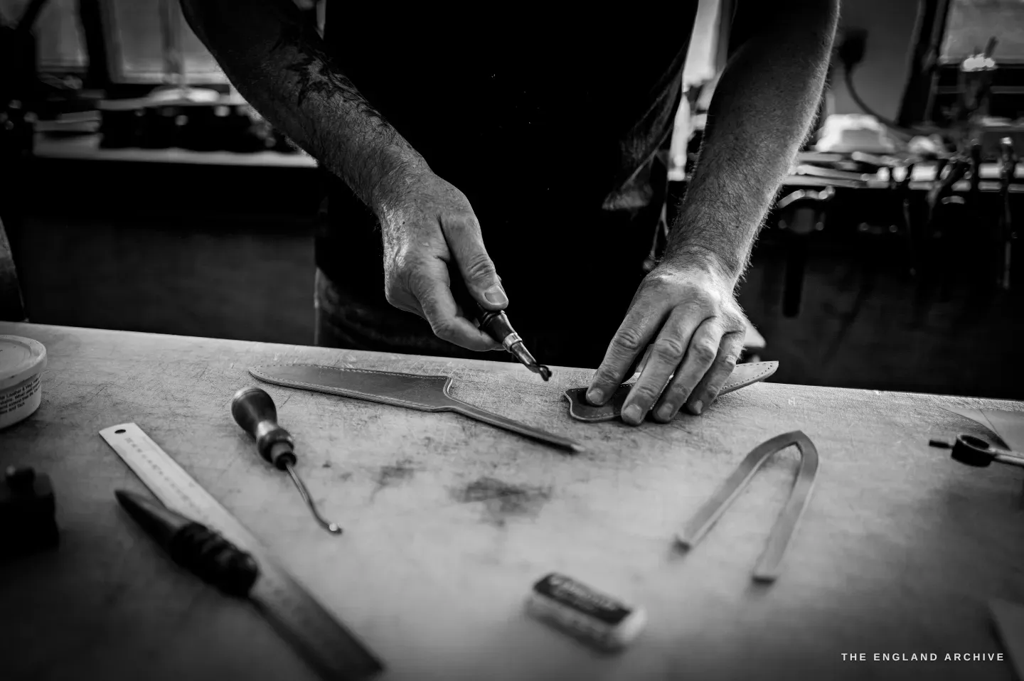 Kevin Wilebore&rsquo;s hands cut into a piece of leather with a round knife on the bench, an awl, ruler and dividers laid out.