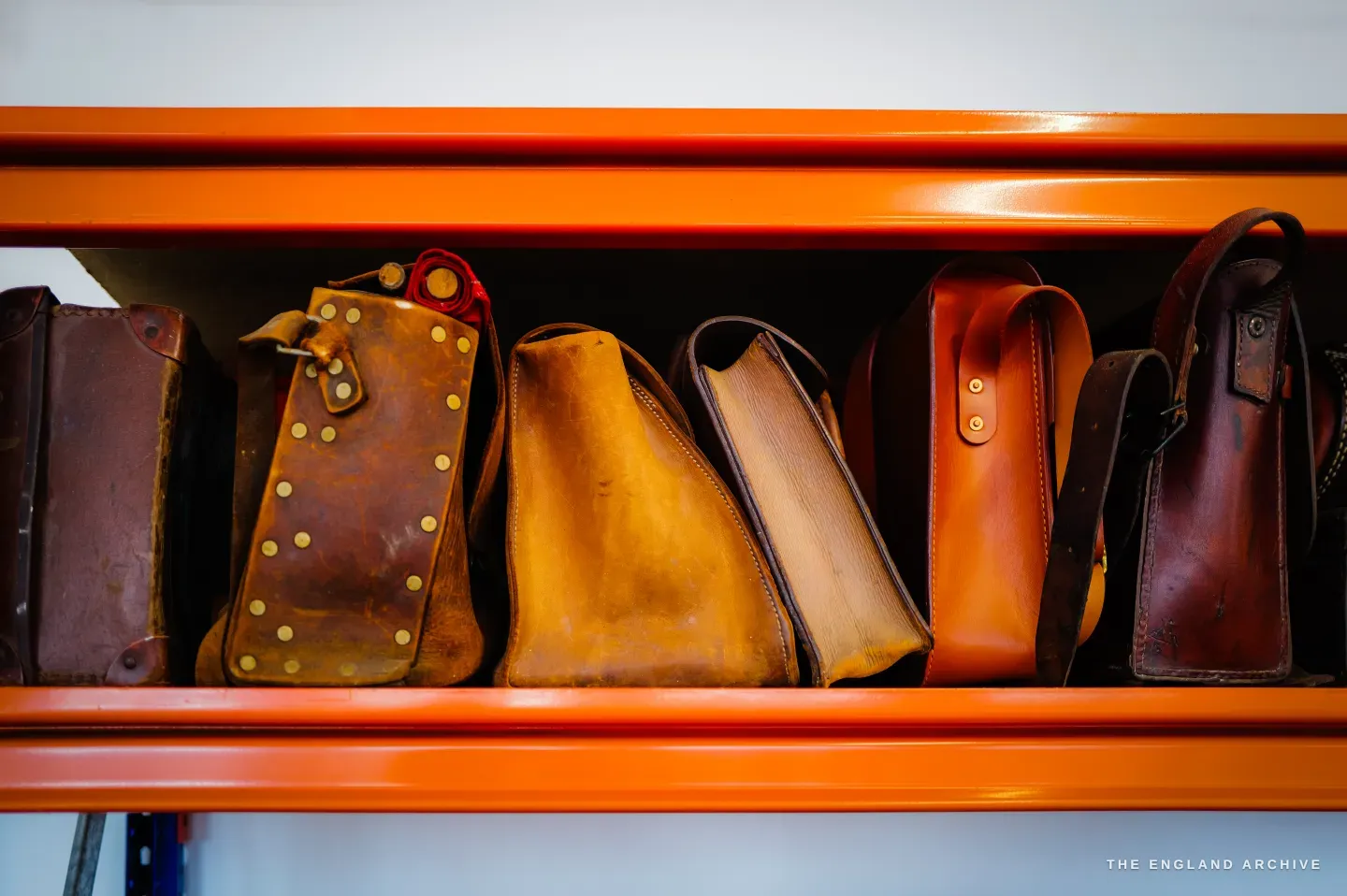 A shelf of finished leather bags and sheaths in tan and cognac on bright orange racking.