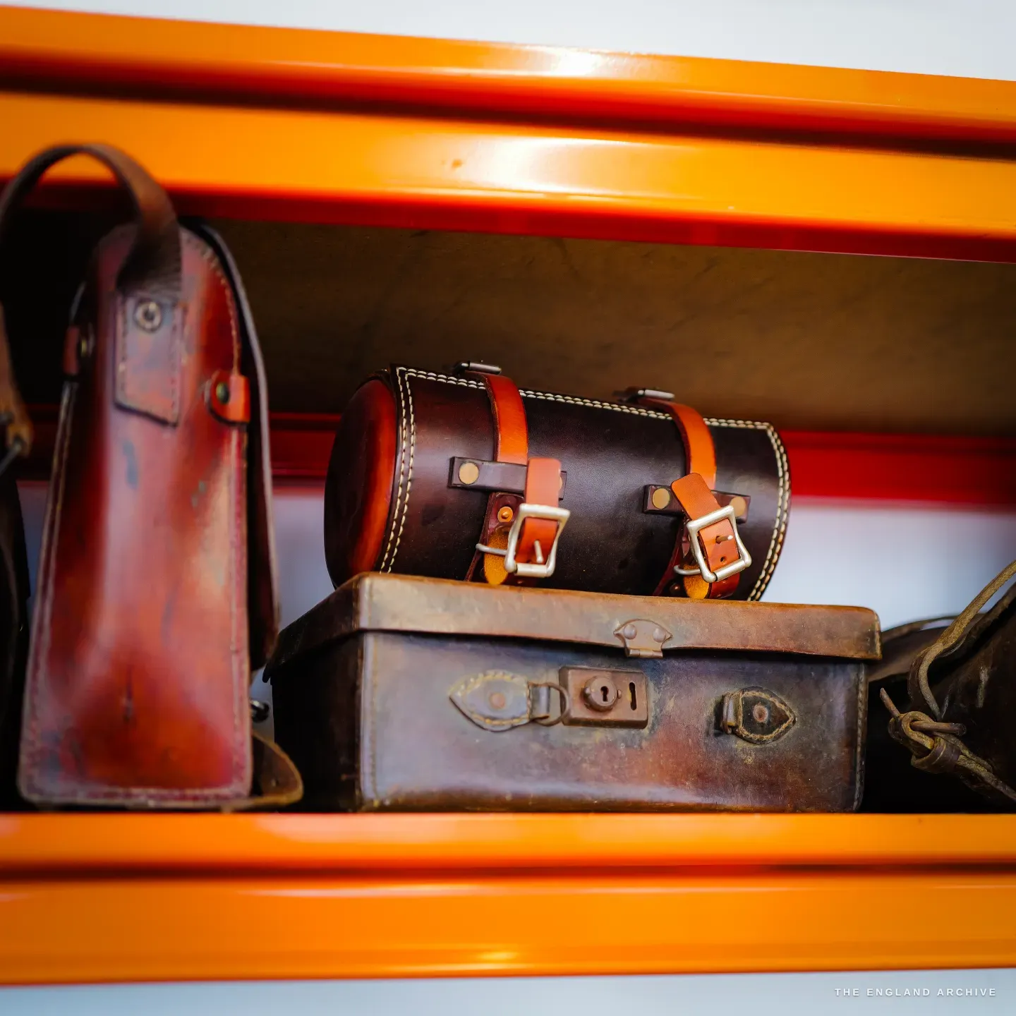 A round leather travel case with buckled straps sits on a shelf beside vintage leather cases on orange racking.