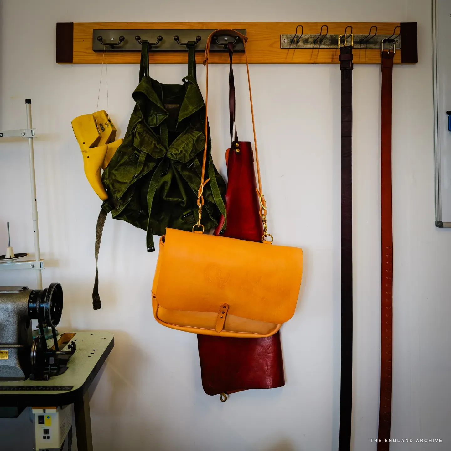 A wall rack hung with Kevin Wilebore&rsquo;s work: an orange leather messenger bag, a red apron, a yellow holster and rows of belts, an industrial sewing machine below.