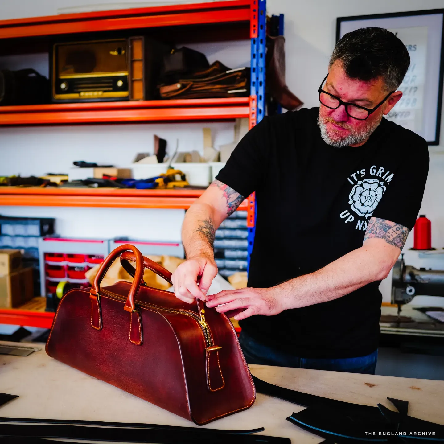 Kevin Wilebore fastens the brass zip of a cognac-leather weekender bag, a vintage radio on the orange shelving behind.