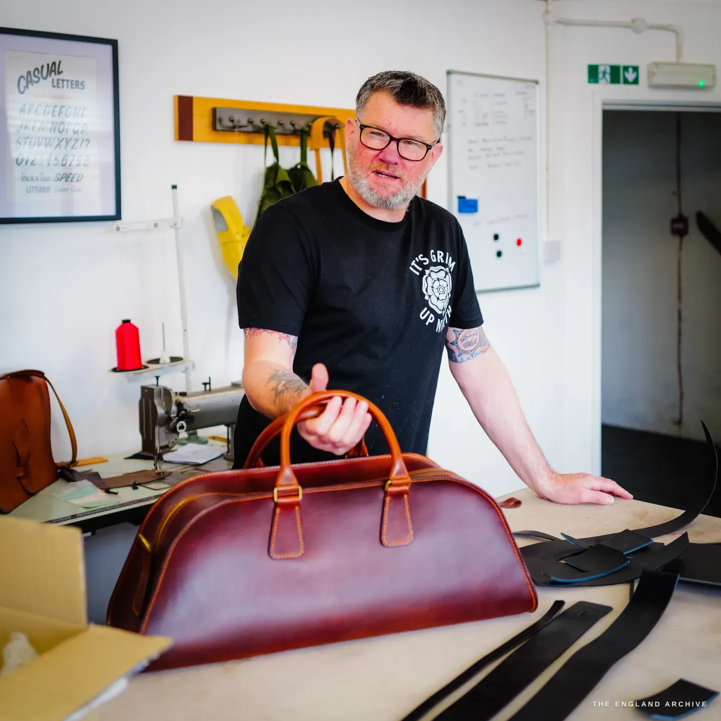 Kevin Wilebore presents the finished cognac weekender bag on the bench, a Casual Letters alphabet poster behind him.