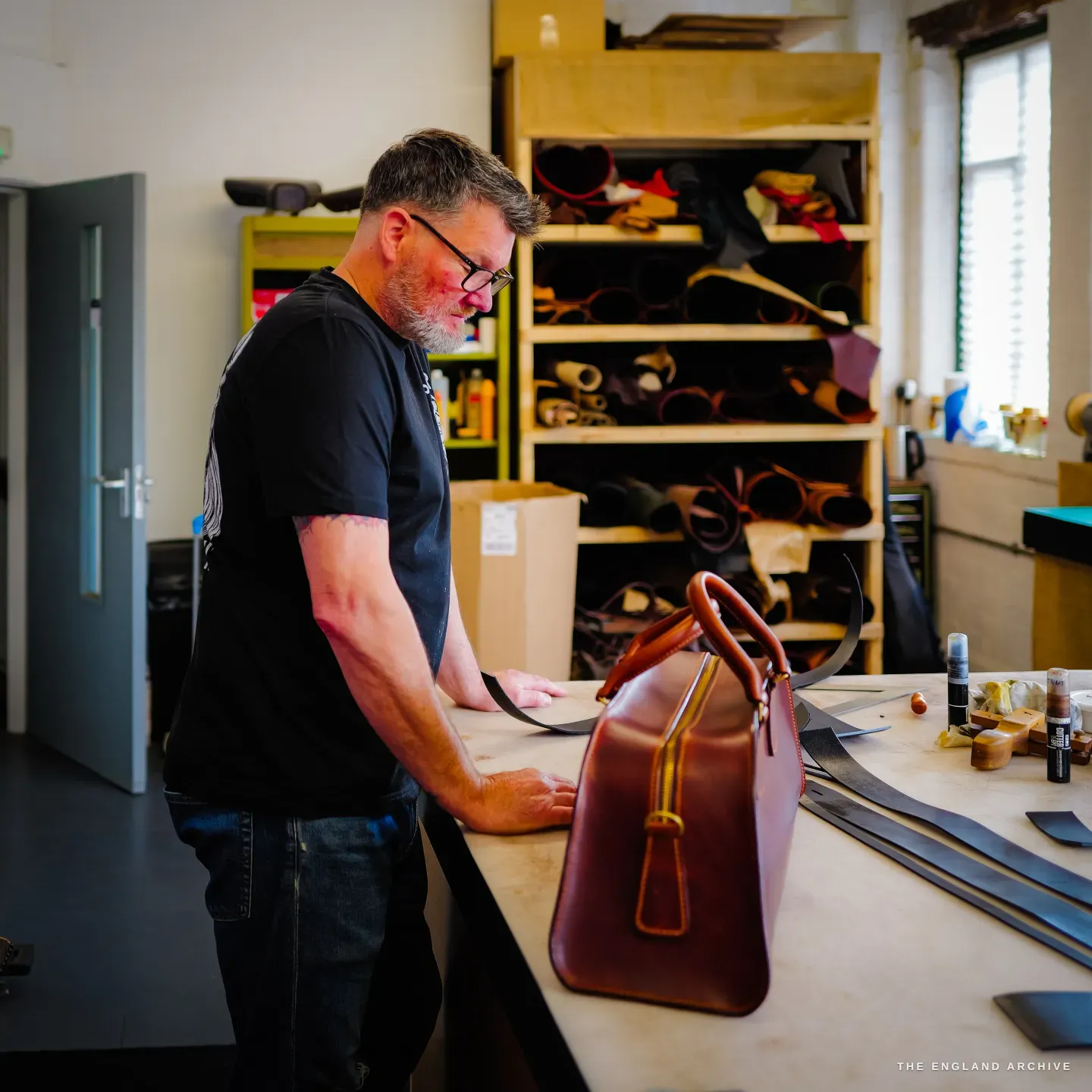 Kevin Wilebore stands looking at the cognac weekender bag on the bench, shelves of hides behind.