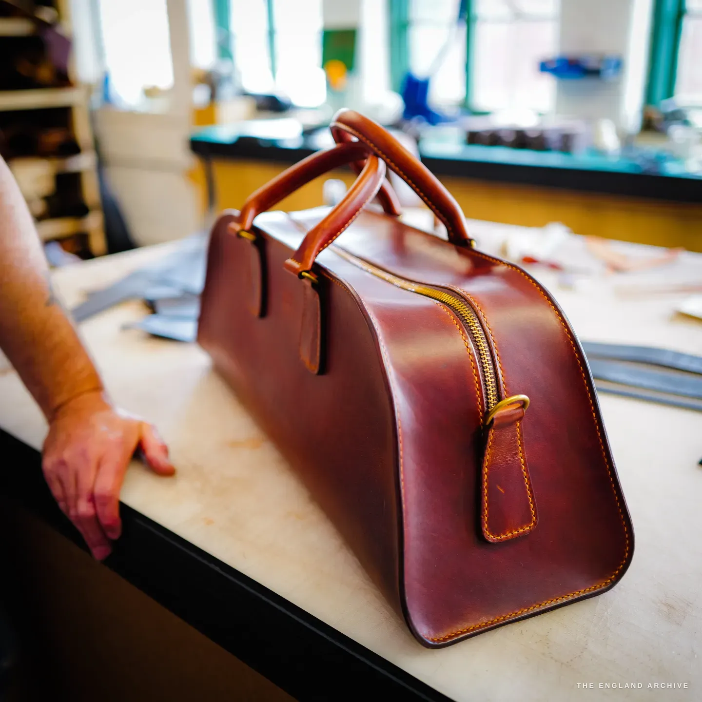 The cognac-leather weekender bag in three-quarter view on the bench, its brass zip and tan handles catching the light.