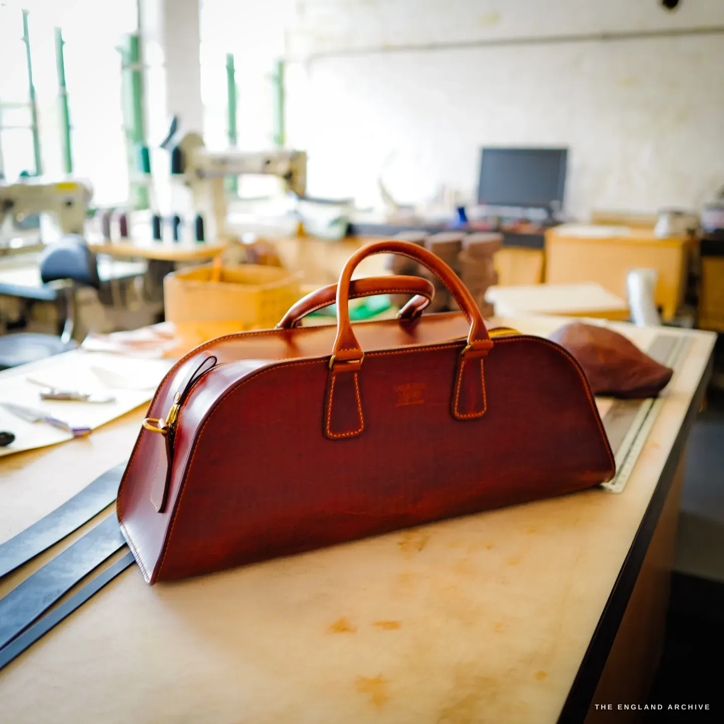 The cognac weekender bag standing on the bench, sewing machines and bright windows behind.
