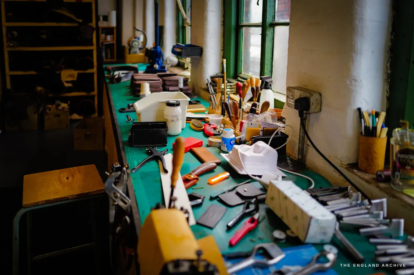Kevin Wilebore&rsquo;s long workbench crowded with leather tools, dyes and offcuts beneath a green-framed window.