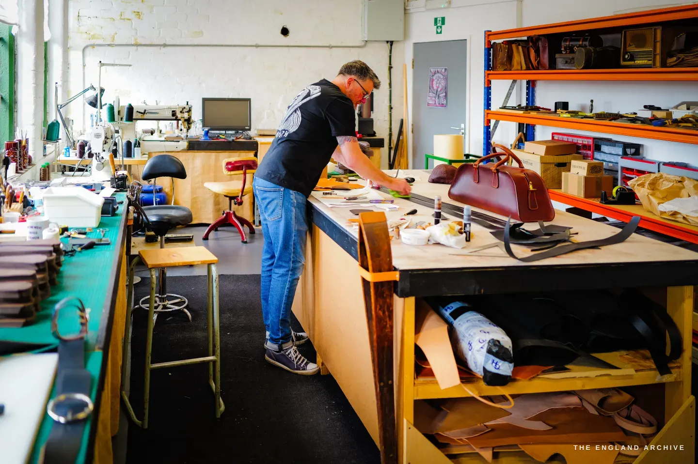 A wide view of Kevin Wilebore&rsquo;s bright, airy workshop, him working at a central bench with orange racking of goods to one side.