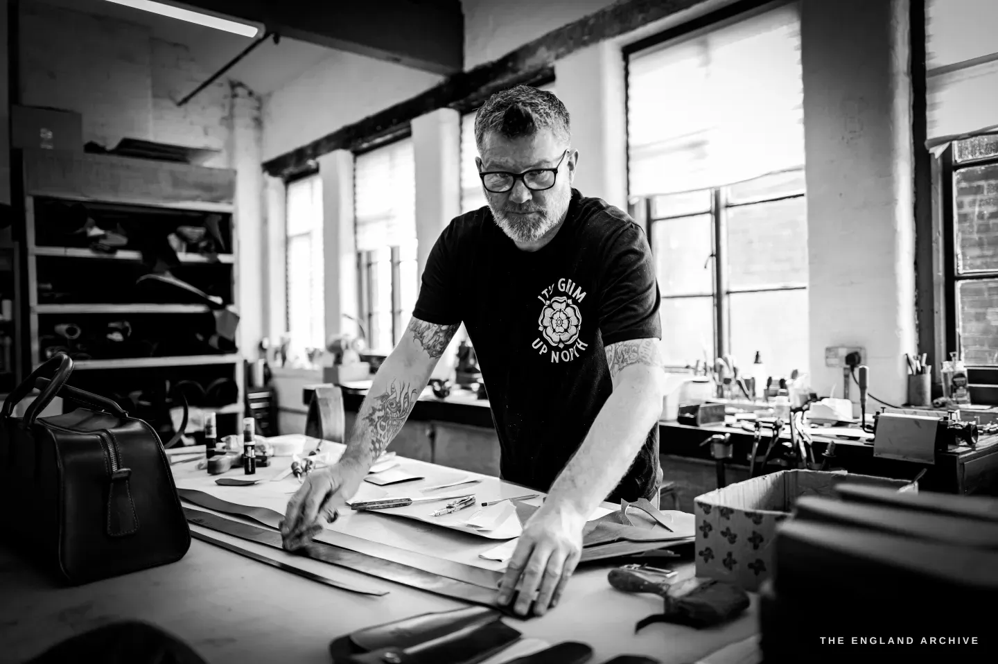 Kevin Wilebore works at the bench beneath the tall windows of the old works, leather and tools spread in front of him.
