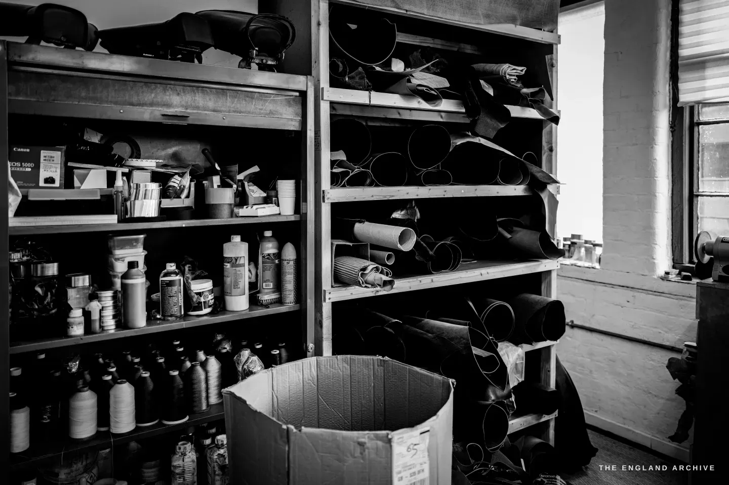 Kevin Wilebore&rsquo;s store: metal shelves of finishing bottles and rolled hides beside a bright window.