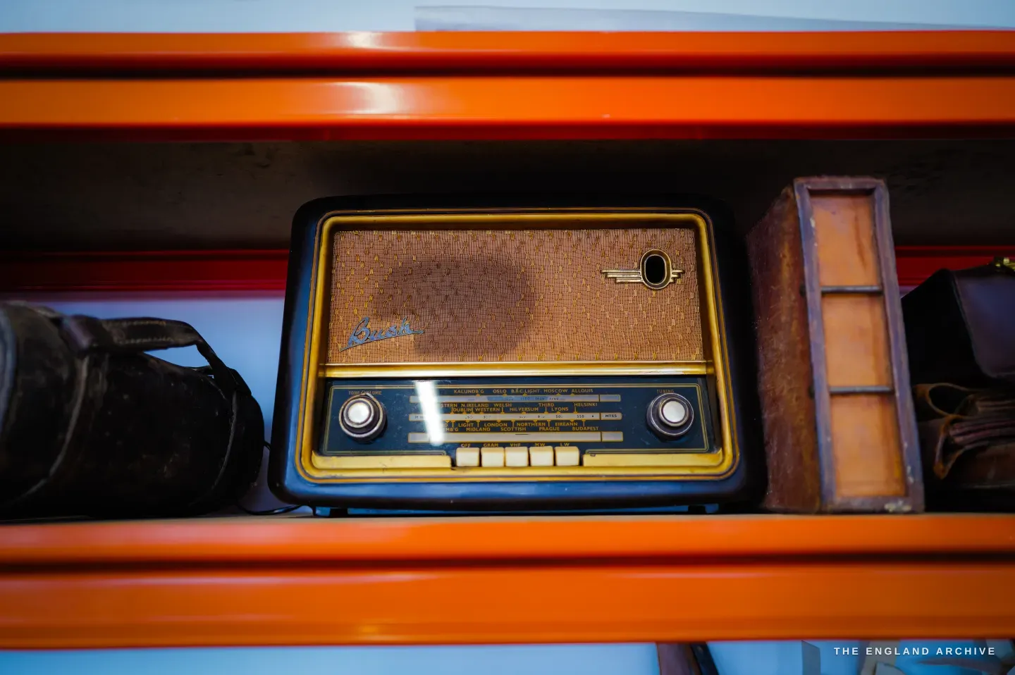 A vintage valve radio sits on the orange shelving among leather cases.