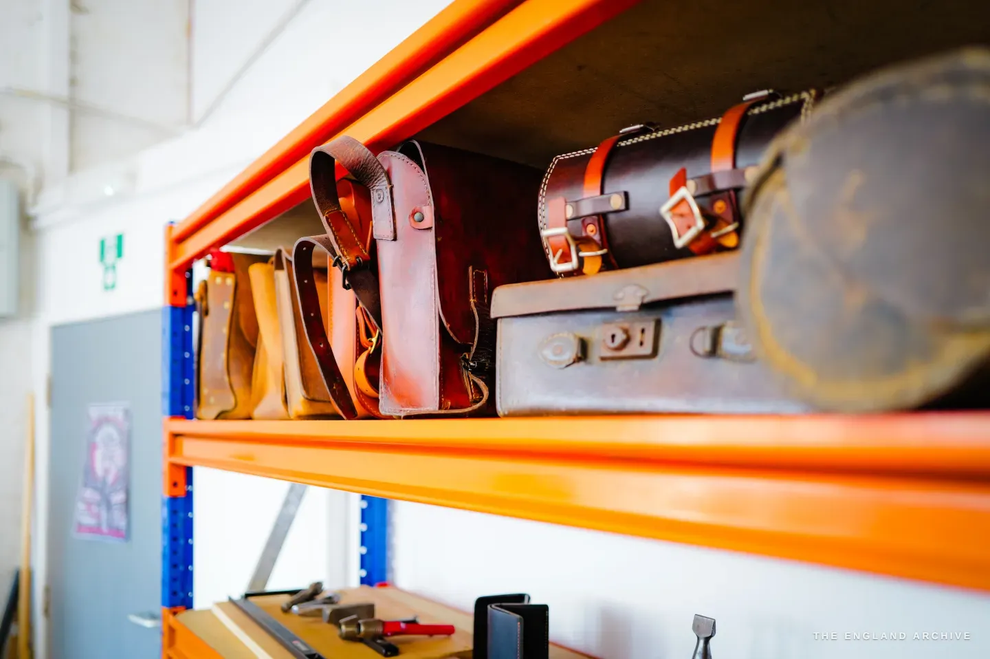 Finished leather goods and vintage cases lined along the bright orange racking - belts, a sheath and a round travel case.