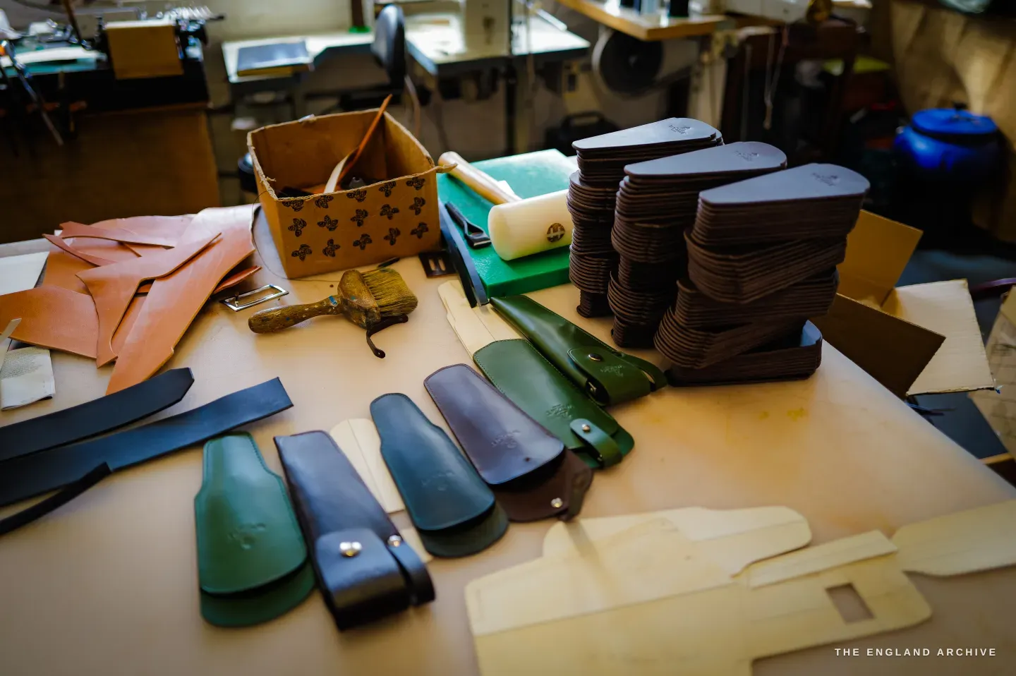 A bench laid with cut leather pouches and sheaths in green, oxblood and tan, with stacks of brown blanks behind.