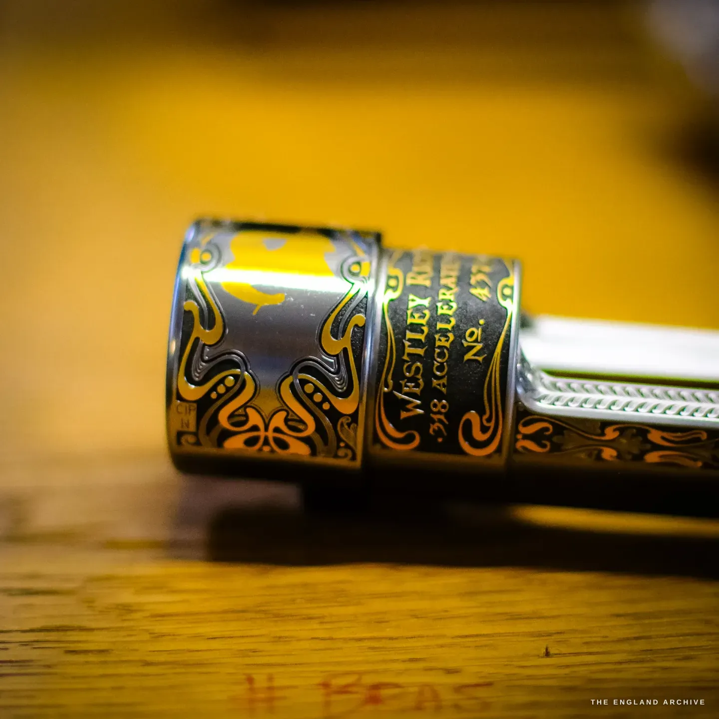 A close view of a gold-inlaid rifle component reading WESTLEY RICHARDS .318 ACCELERATED, gold scrollwork and serpents cut and inlaid into the blued steel on a warm ground.