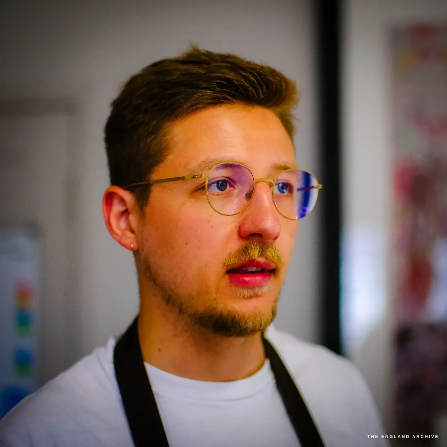 A close portrait of Léo Lambert, a man in his late twenties with short hair, a moustache and round glasses, in a white t-shirt and apron strap, looking a little off to the side in the studio.