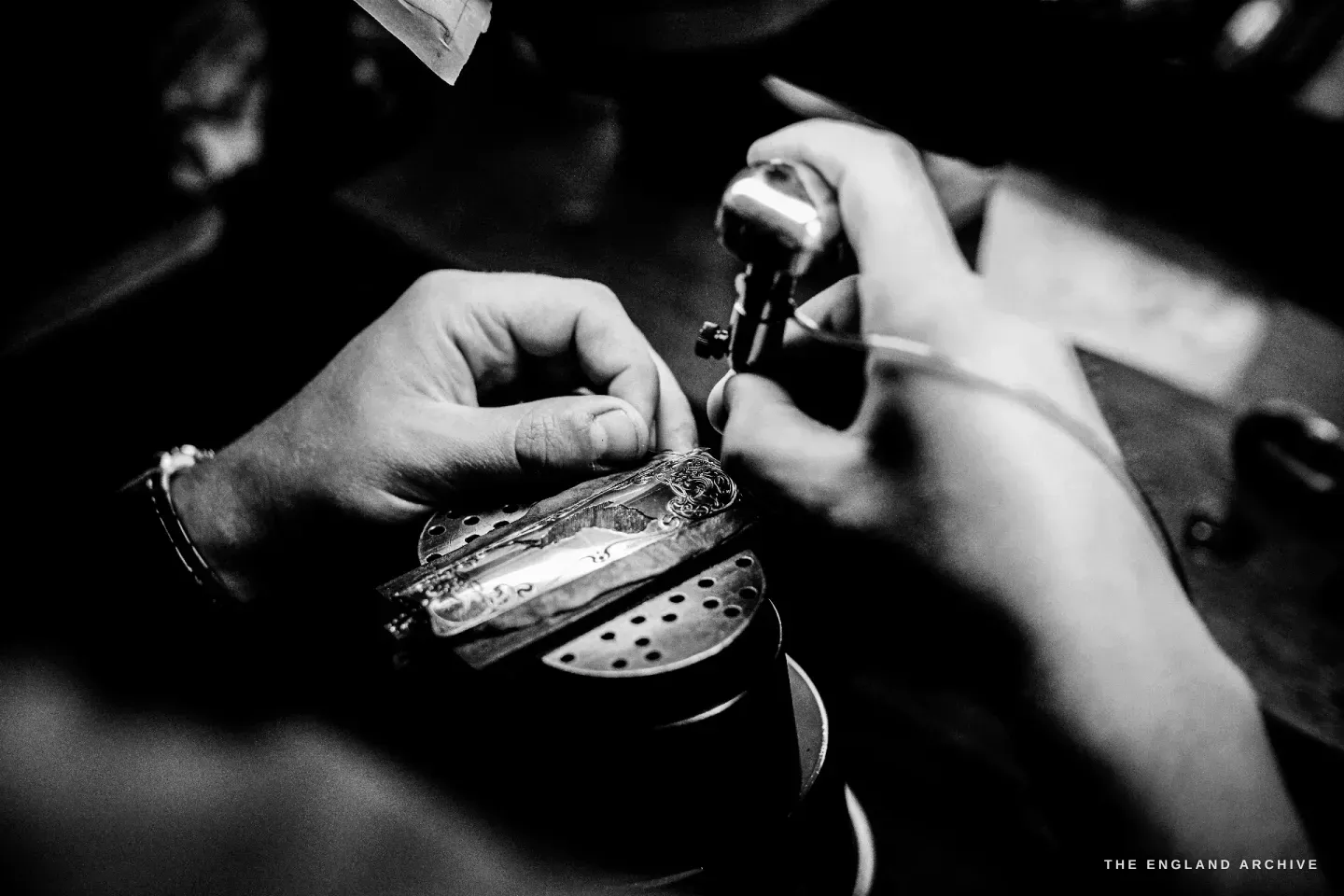 Hands hold a small bright gun component and a graver at the bench, mid-cut, in close focus under the lamp.